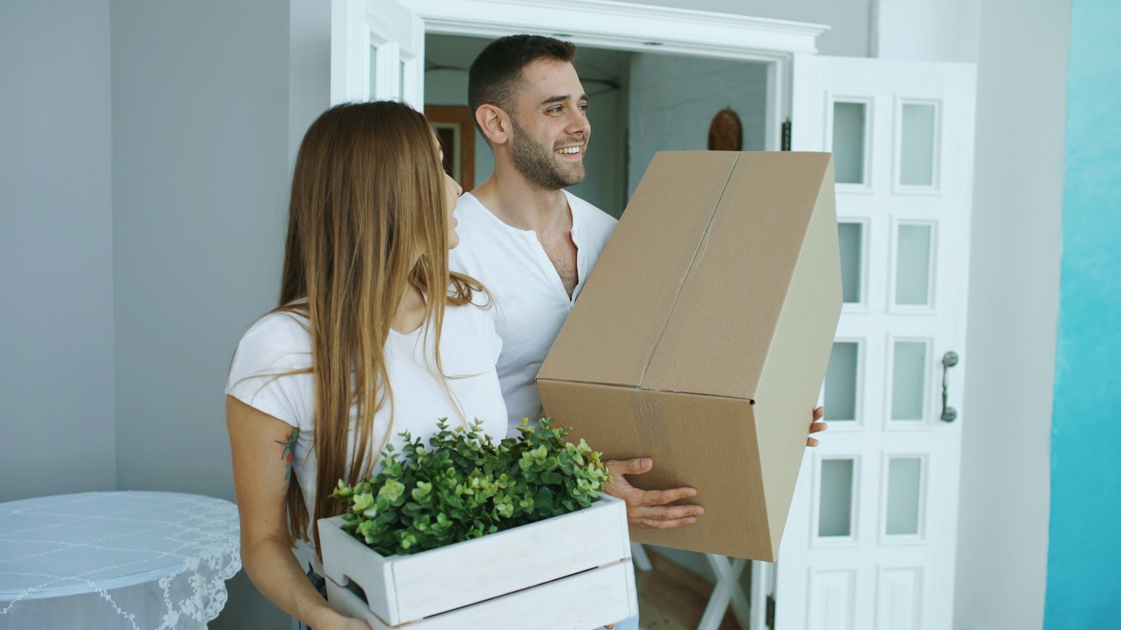 A couple carrying moving boxes and a plant into their new home